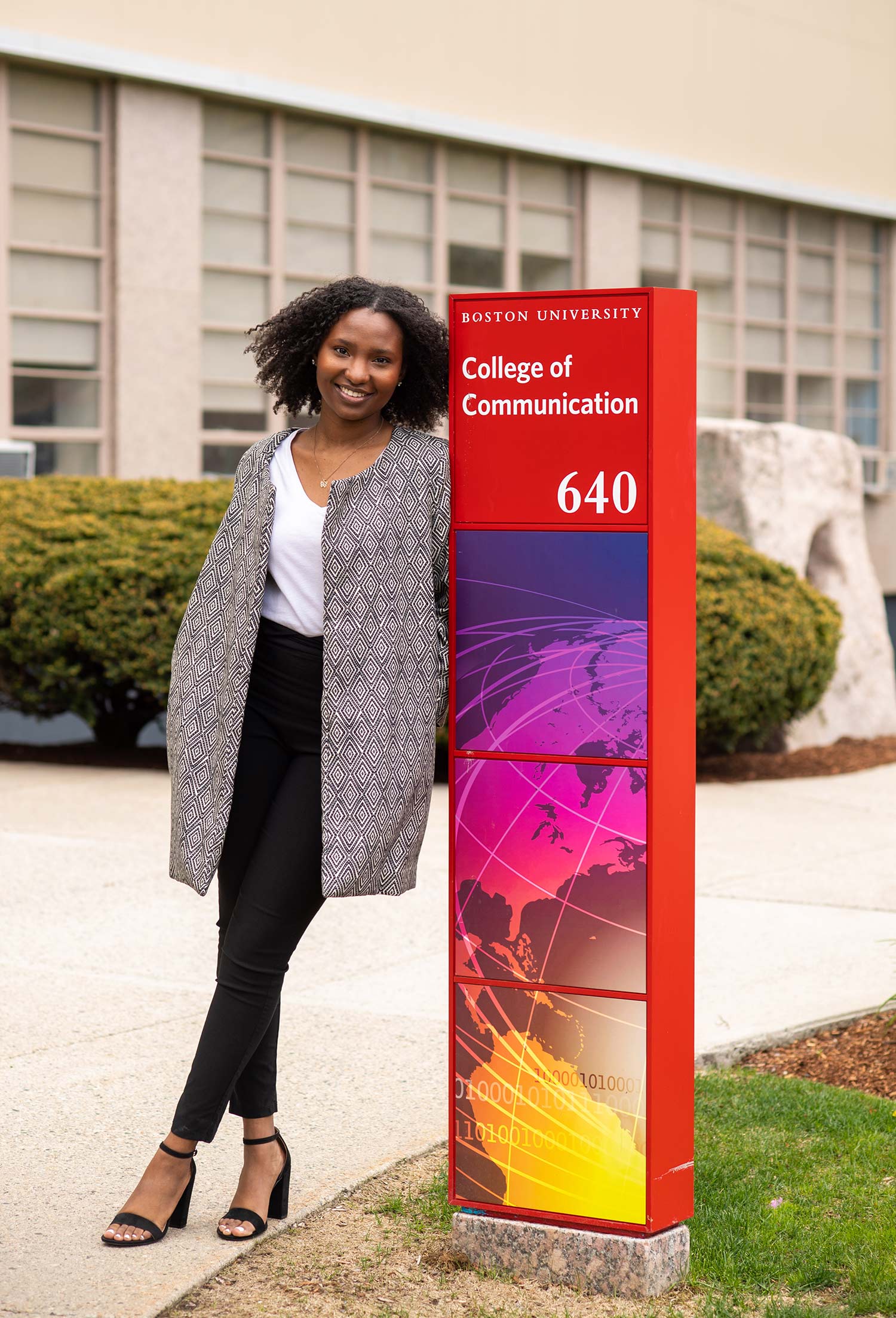 Portrait of Shadae Leslie (COM’20) in front of COM May 12, 2020. Leslie leans against a red "Boston University College of Communication" sign.
