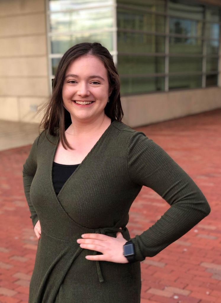 Portrait of Sarah Bickford (CAS’20, wearing a green dress and poses in front of a building on a brick sidewalk.