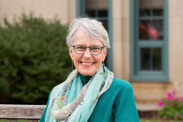 Photo of Dean Moore sitting on a bench in a scarf and teal shirt. Flowers and a grass patch are seen behind her.