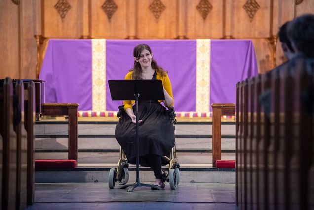 Elizabeth Foster (STH’22), who has a condition that makes prolonged standing difficult, preaching from her walker chair during a Marsh Chapel service dedicated to those with disabilities. An altar with a purple cloth is seen behind her.