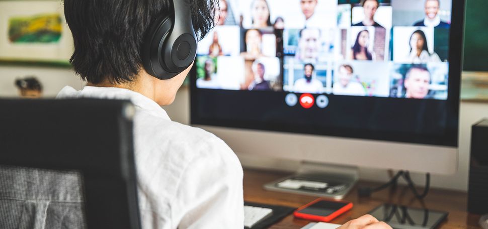 Photo of a student with headphones looking at a desktop screen with a video chat call is seen. The student's hand is on the mouse.