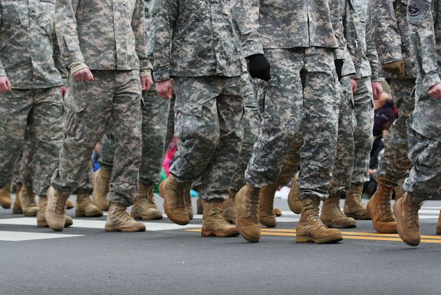 Image of soldiers dressed in army camouflage in an army parade. In the shot, only the road, army boots, and lower half of the soldiers are seen.
