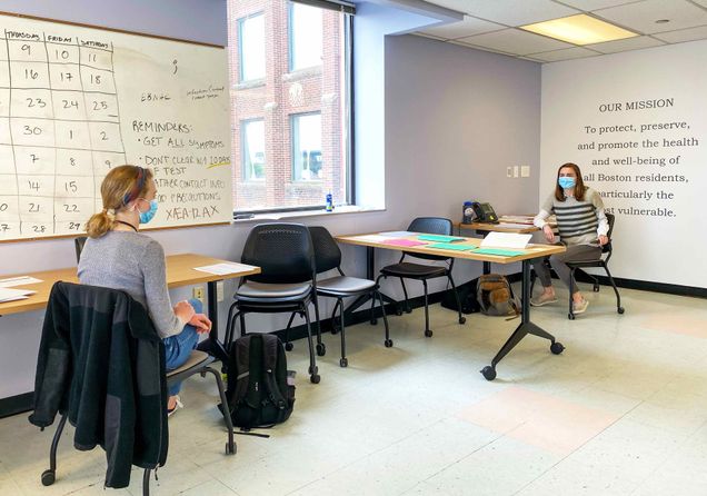 Jessica Schueler (SAR'21) and Natalia Kelley (SAR'22) volunteer as contact tracers as part of the city of Boston's coronavirus pandemic response. They are seen here, socially distant, at desks in an office. A white board and mission statement are seen on the walls.