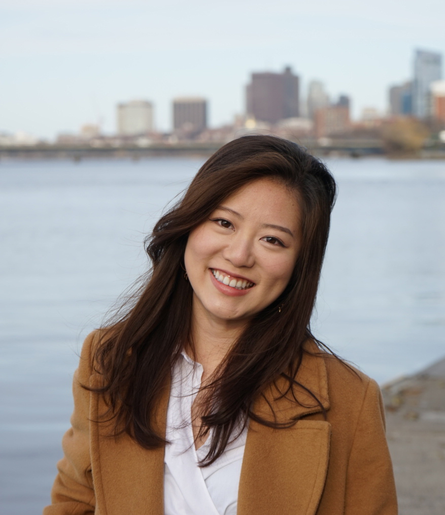 Portrait of Rinka Murakami (SAR'20, MPH'21 in a light brown blazer. A blurry city skyline and water is seen in the background.