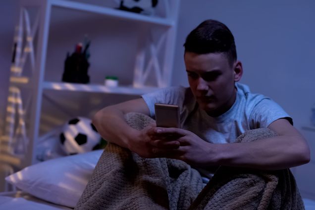A photo of a person sitting on a bed in pajamas looking at their phone with an upset expression on their face