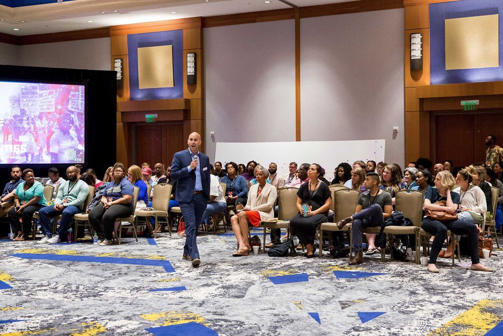 Patrick Reyes speaking at at a workshop. Reyes wears a blue suit and walks in front of participants seated in chairs.