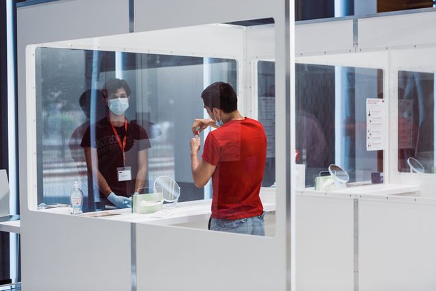 Photo of Covid-19 testing lab at Kilachand Center. A young person in a red t-shirt in a testing both takes a COVID testing swab out of a tube as a testing site worker looks on.