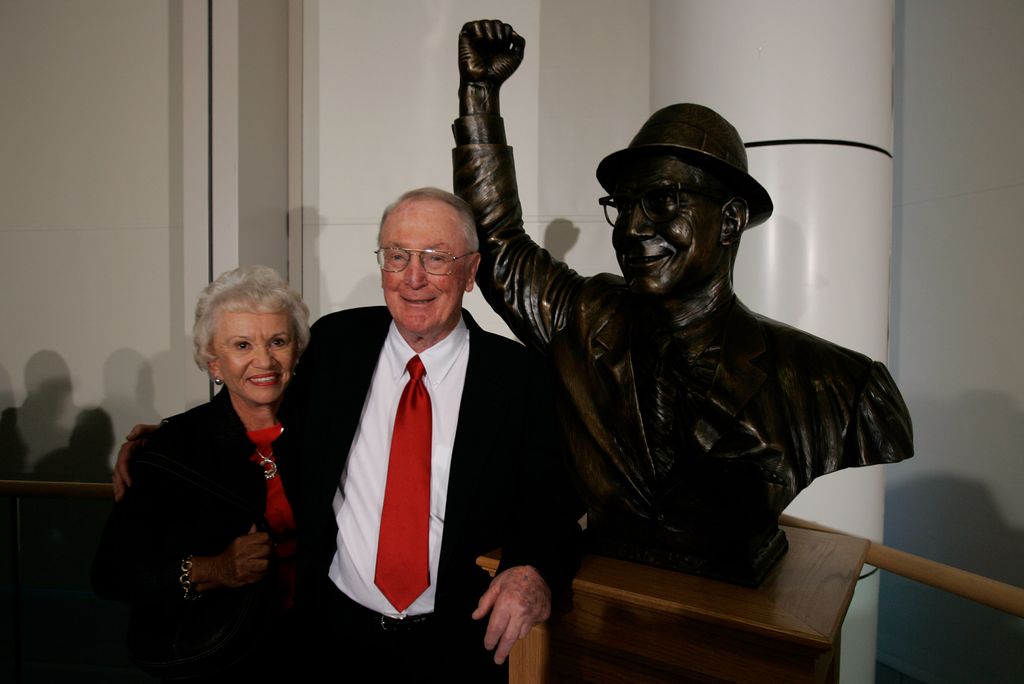 Photo of Jack Kelley with his wife, Ginny, at Agganis Arena, next to a bronze statue of Kelley with his hand in the air.