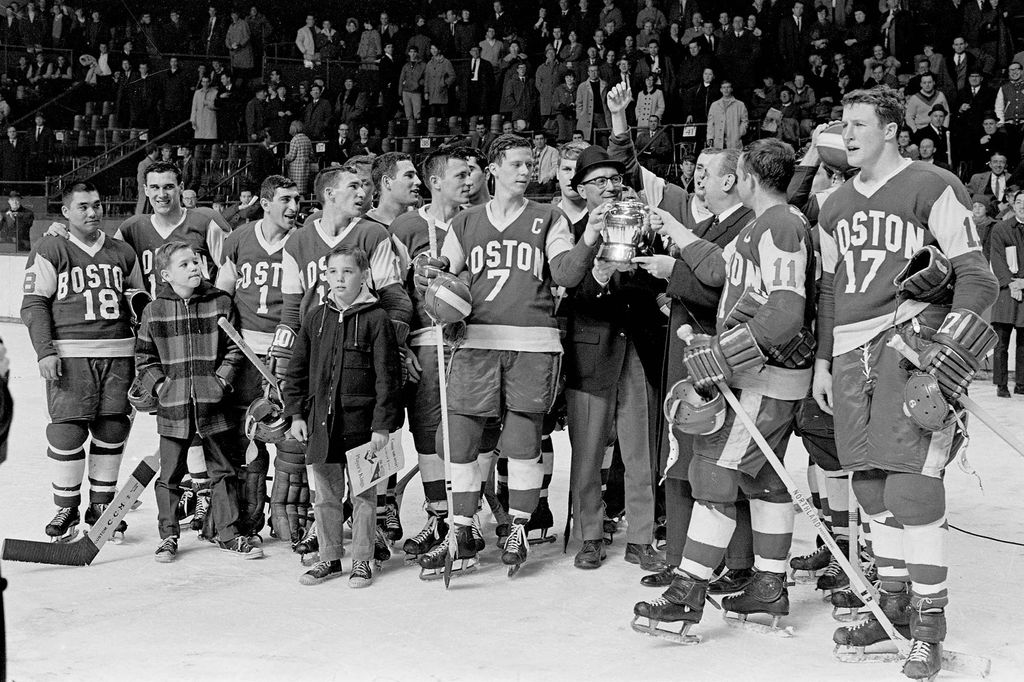 Black and white photo of men's hockey team on the ice after Boston University defeats Northeastern 4-0 in the Beanpot Tournament final game. BU Players include Herb Wakabayashi #18, Jim Quinn #7, Pete McLachlan #11, Bill Hinch #17.  David Kelley on ice with his dad, Coach Jack Kelley.