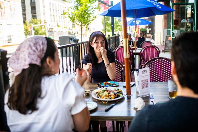 Photo of student enjoying nachos with her friends Caroline DeHaven (SHA 23) and Jason Wilmot (SHA 21) outside at Sunset Cantina on September 8, 2020. Student smiles across the table from her friends; the group sits outside on the patio, a sidewalk is seen to student's right.