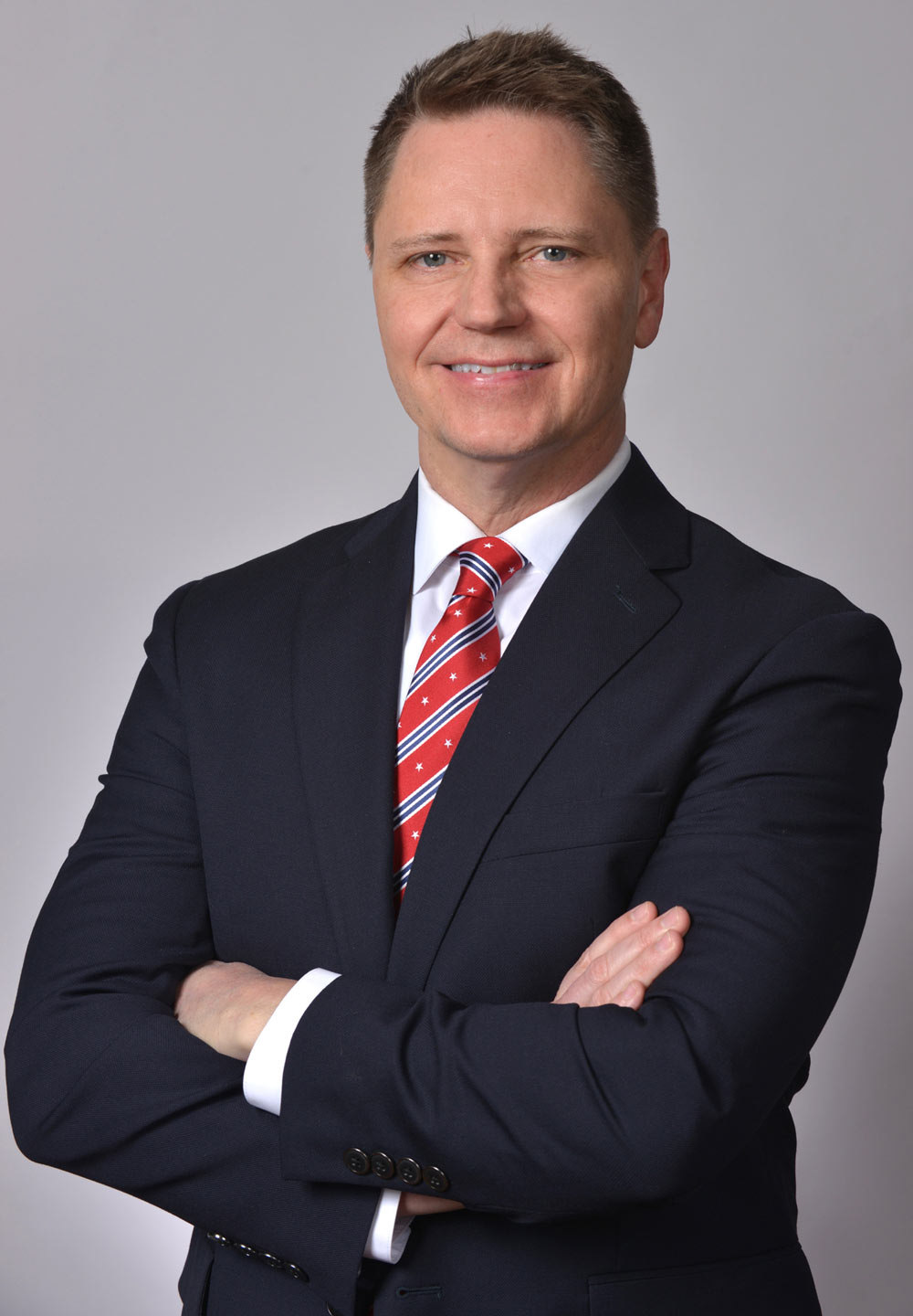 Portrait of John Paul Moran (Questrom’05) who is seeking his first term in office, in the Massachusetts 6th District. She wears a blue suit and red and blue stripped tie, stands smiling with his arms crossed.