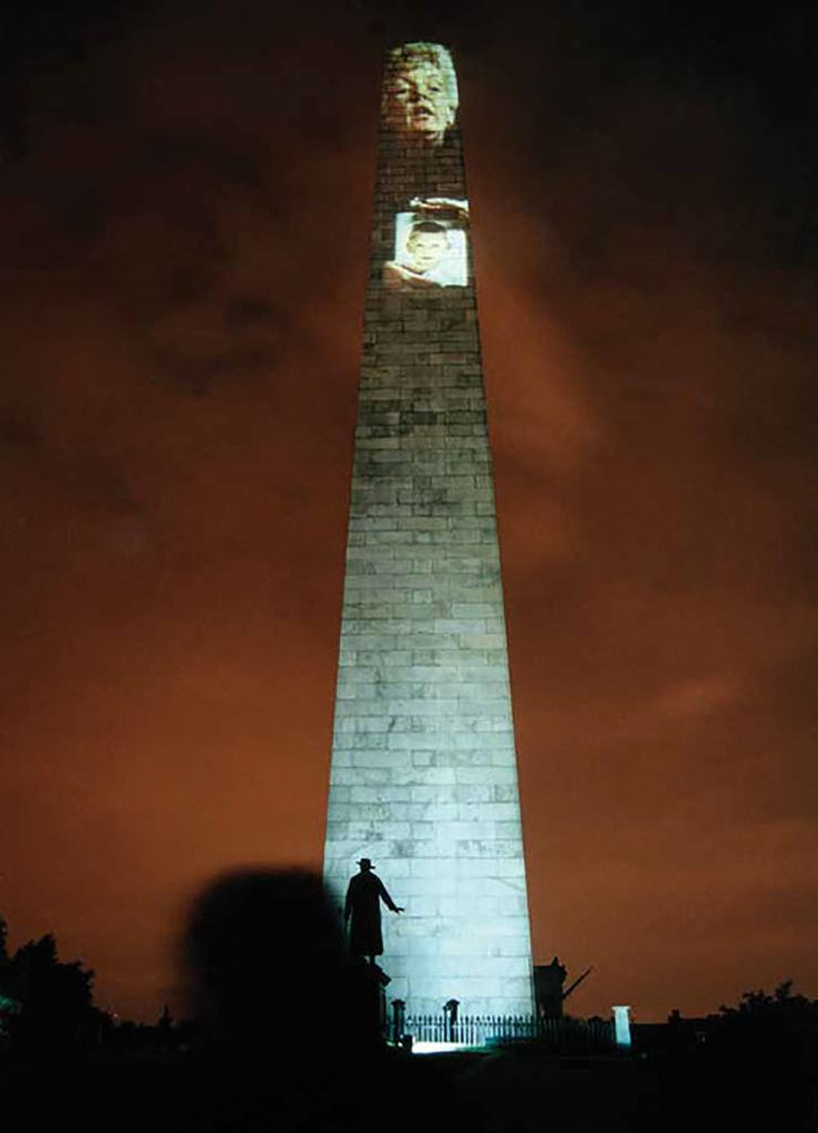 Photo of Krzysztof Wodiczko's artistic projections on the Bunker Hill Monument in Charleston which occured in 1998. The artist transformed a historical monument devoted to freedom fighters into a monument of contemporary heroes trying to fight violence. On top of the huge obelisk were projected images of the faces of people recounting their experiences, mothers of murdered children, or young people who had lost their brothers or sisters. Shown from the waist up, they held candles or portraits of their dead loved ones, the huge images