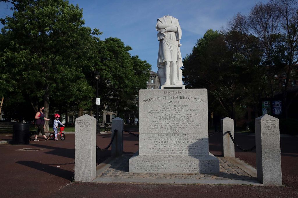 Photo of the statue of Christopher Columbus in Christopher Columbus Park in the North End neighborhood of Boston, MA on on June 10, 2020 after the statue was beheaded overnight. A blue sky and trees are seen behind the statue, as well as people walking through the park.