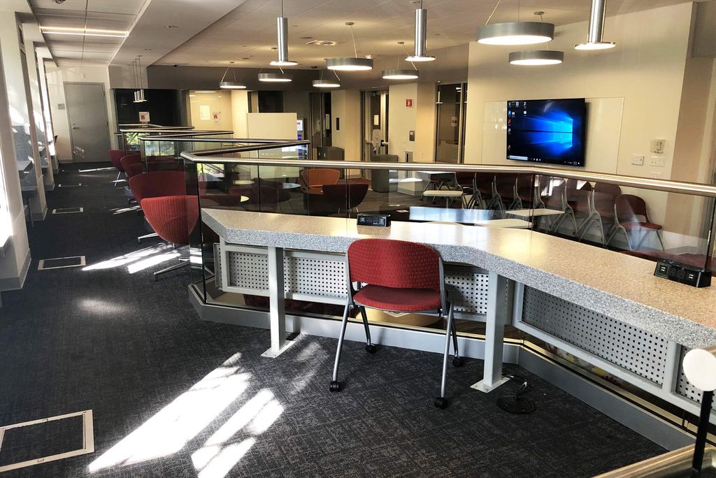 Photo of the Think Tank in Room 105 at 725 Comm Ave. Red chairs are place along a long table, a TV and additional seating are seen in the background. Available spaces are designated for a specific number of people, from one to 25.