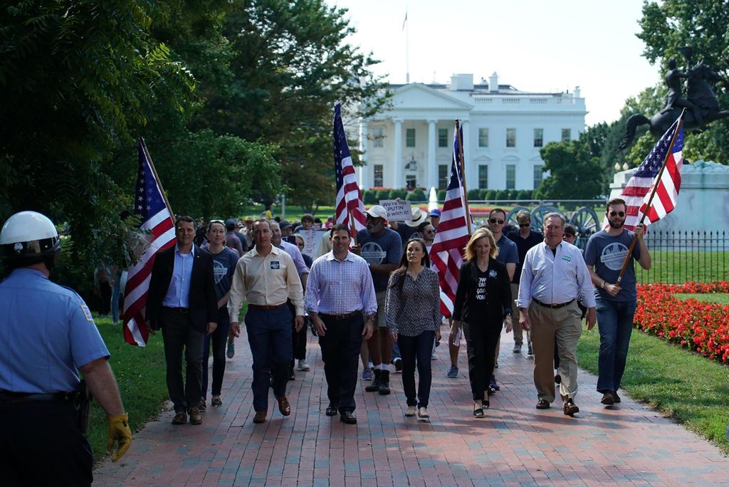 Photo of Gina Ortiz Jones (CAS’03, GRS’03) (center) marching outside the White House in 2017 with a group working to increase veteran representation in Congress. Ortiz wears a black and white tripped blouse and looks ahead with a group of mostly men around her. The white house and a statue are seen in the background.