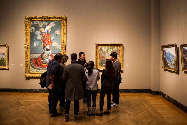 Photo of a group of students gathered in the MFA in 2018. Behind the students are two large works of art in ornate frames. A semi -circle of students listen as one student presents their work of choice.