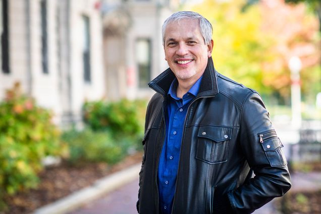 Portrait of Eugenio Menegon, College of Arts & Sciences associate professor of Chinese, wearing a bright blue button down and black leather jacket. He smiles and a fuzzy background with a building and fall trees are seen behind him.
