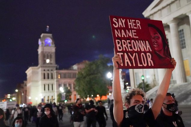Photo of a protestor holding a red sign that reads "SAY HER NAME BREONNA TAYLOR" during a September rally in Louisville, Kentucky. The protestor has a black power fist on their face mask, a crowd of people and a tall white building are seen behind them.