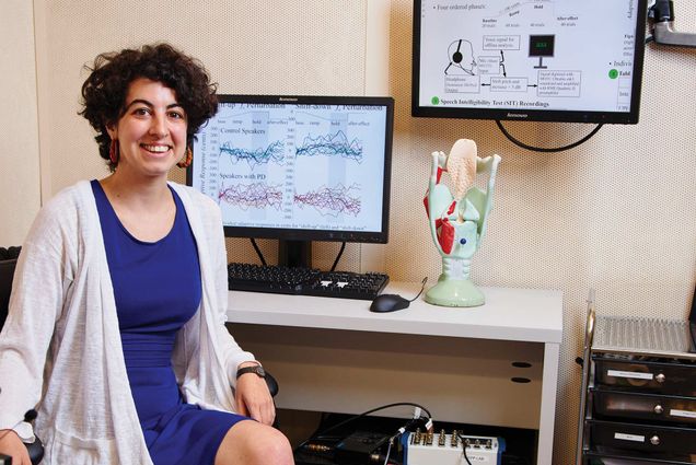 Photo of Defne Abur in a white sweater and bright blue dress and red shoes sitting at her desk in front of two computer screens. On her desk is a medical model of the lungs.