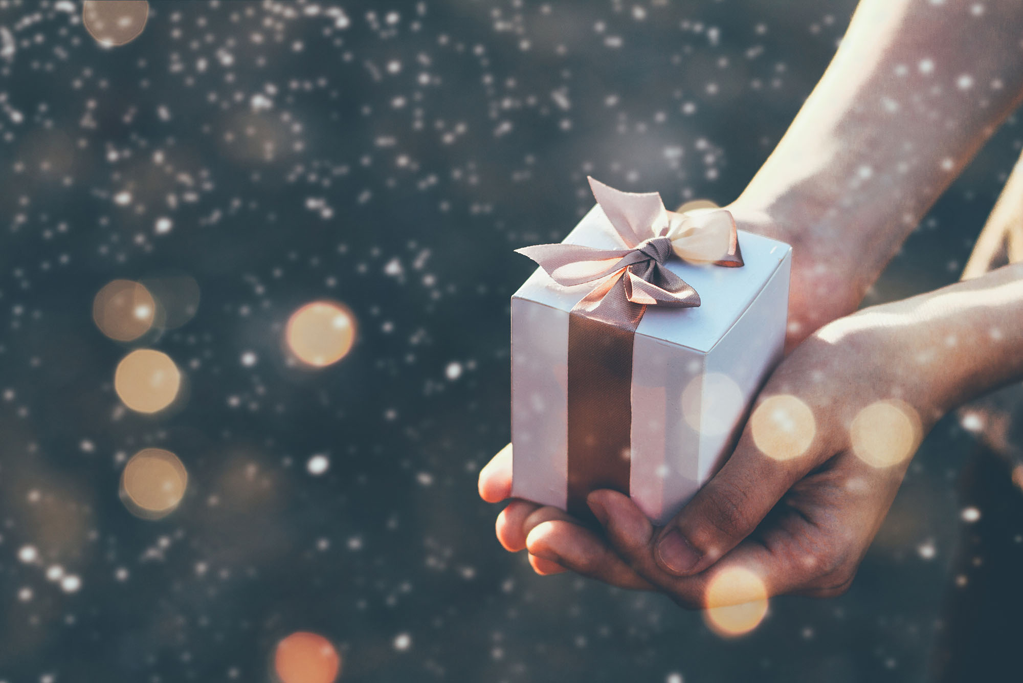Photo of a person holding a small white box with a golden ribbon. In the background it appears to be snowing.