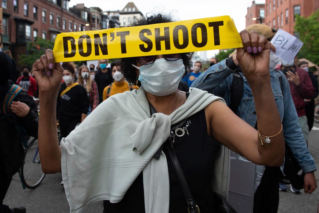 A photo of a masked protestor holding up yellow crime scene tape that reads "Don't Shoot"