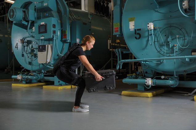A woman in all black workout gear squats as she wears a soft exosuit developed by researchers at Sargent that helps mobility and movement for people suffering from lower back pain, aiding them in activities such as heavy lifting. Behind her large blue industrial vats are seen.