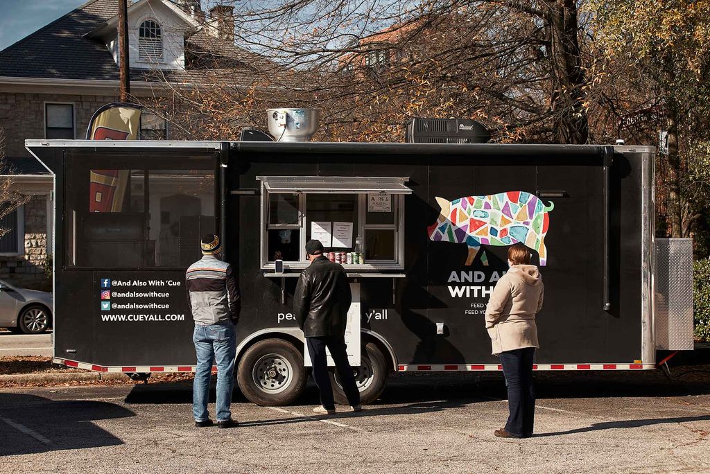 Photo of three people of various ages, wearing jackets, waiting outside of David With’s BBQ trailer, either for their food or to order. The trailer features a colorful, mosaic logo in the shape of a pig.