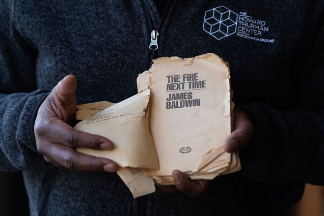 Photo of Dean Elmore holding a tattered copy of "The Fire Next Time by James Baldwin. The photo is a tight shot on Elmore's hands as he opens to the book's yellowed title page.