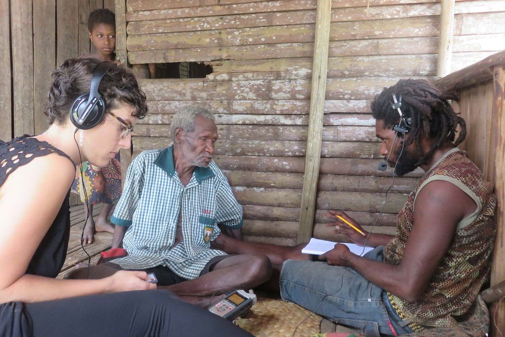Photo of Kate Lindsey recording and listening to two people in Papua New Guinea as they speak. A small child looks on from the door way. Lindsey wears headphones and looks down at a recorder as her subject speaks into the microphone of a headset. 