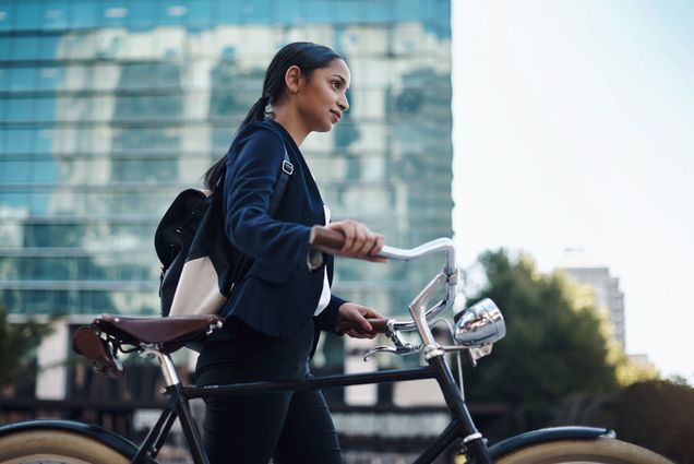 A photo of a woman wearing a business suit walking a bike. A cityscape is visible behind her.