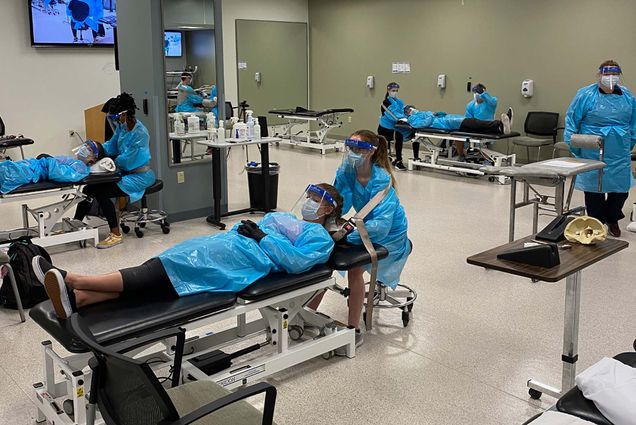 Photo of students in Sargent’s occupational therapy program in full blue PPE gowns, face shields and masks, during a training session. One student lays on the bed while the other attends to mock-patient.