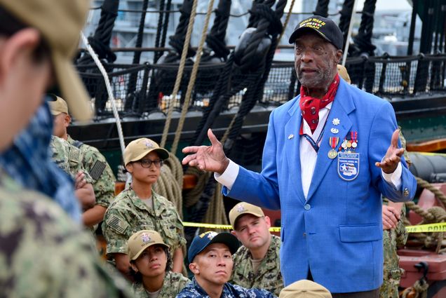 Photo of Retired U.S. Air Force Lt. Col. Enoch Woodhouse in a blue suit jacket, baseball cap, and red neck-tie with his hands extended towards a crowd of chief petty officer selects aboard USS Constitution, who wear camo and tan baseball caps.