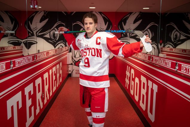A photo of Logan Cockerill holding his hockey stick over his shoulders in the BU hockey dressing room