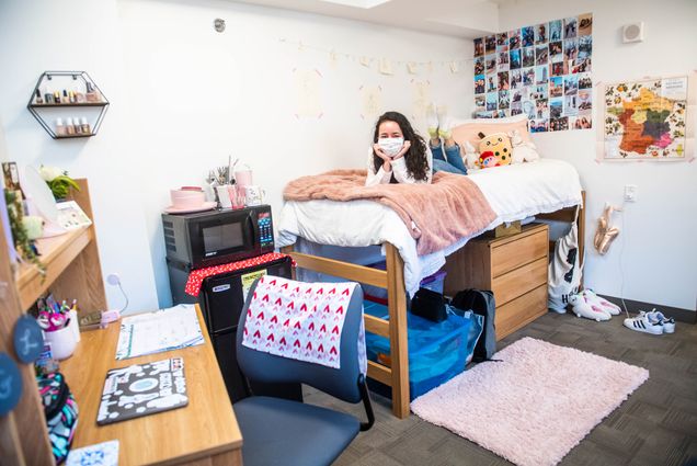 A photo of Léa Namouni sitting on her bed in her dormroom. The room is decorated with a map of France, photo collage, a nail polish shelf, and a fuzzy pink rug.