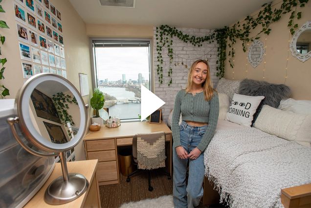 Photo of Casey Evans (COM’21) standing in her StuViII quad with a million-dollar view of Boston’s skyline in the window at the back of the room. The dorm room is beautifully decorated with hanging plants, a wall of photos, and a bed with many pillows, one of which reads “good morning.”