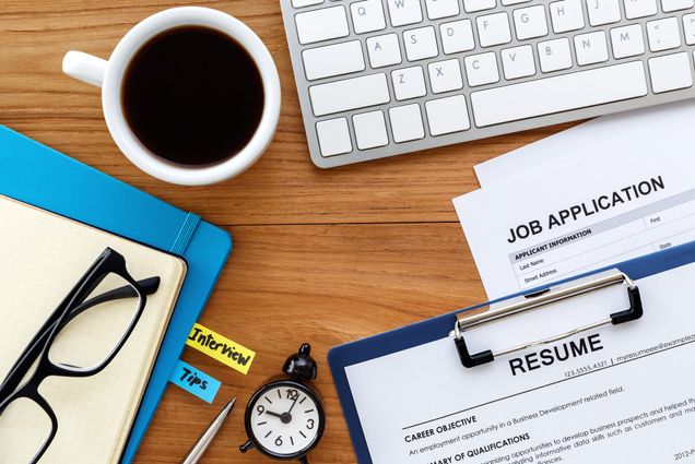 A photo of a desk cluttered with job applications, resumes, a keyboard, a pair of glasses, and a cup of coffee among other things.
