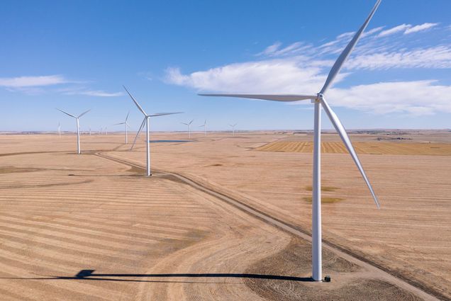 Photo of large white wind turbines in a dry, flat landscape. A blue sky with small clouds are seen.