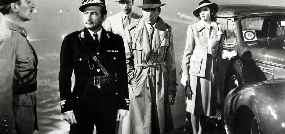 Black and white still from the film “卡萨布兰卡.” In the culminating scene, Jean del Val, Claude Rains, Paul Henreid, Humphrey Bogart, and Ingrid Bergman are seen. The men wear trench coats and hats, and the officers wear uniforms.