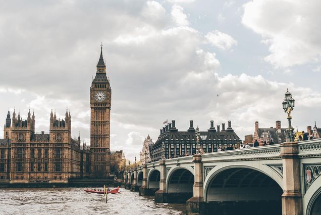 Photo taken from across the Thames River of Big Ben with an ornate bridge on the right; the heads of people walking over the bridge are seen. In the river, a small red boat with people moves to the left; the sky is grayish and cloudy.