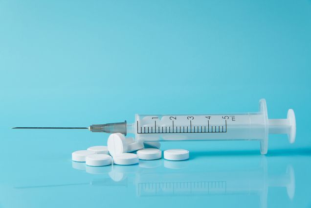 Photo of a pile of white, circular pills with a syringe resting on top; the background is completely light blue. Semaglutide can be administered in both forms.
