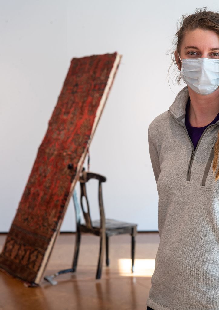 Photo of Hannah Minife (MFA’21) standing in front of part of her sculpture, “Three Doors.” She wears a gray pullover and blue face mask. Her sculpture features a door covered with an elaborate textile pattern, balancing against a chair, with an axe resting on the ground.
