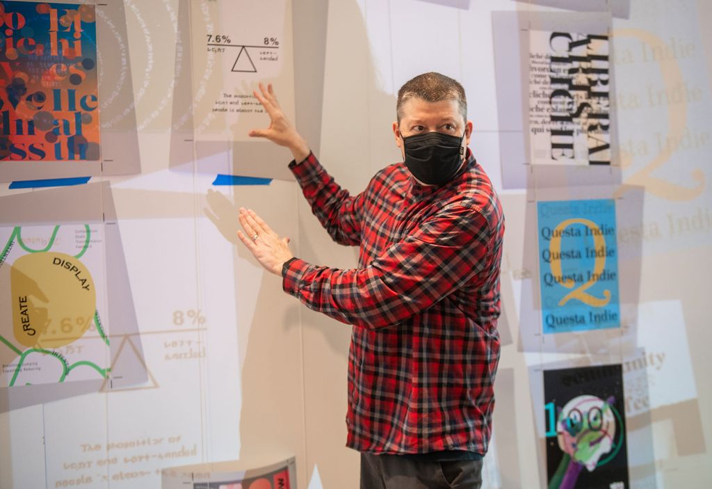 Photo of Christopher Sleboda, in a red and green plaid shirt, in front of the “Scale Wall” in the Stone Gallery, pointing with both hands towards the wall.