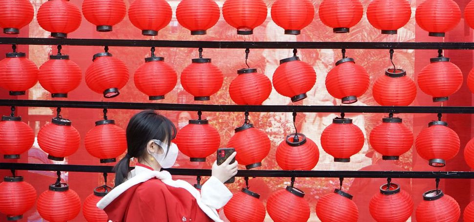 Photo called “New Year from Anywhere” in which a young girl holds up her phone towards a wall of red lanterns; she wears a red coat with an intricate design.