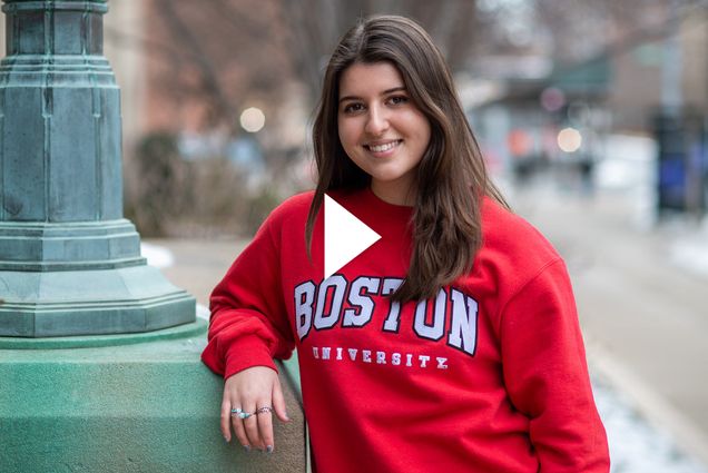 Photo of Vlogger Anna Malygin (Questrom'23), smiling, with her hand resting on the base of a green lamp post. She wears a bright red Boston University Sweatshirt, and has her hair down. Video play button is overlaid.