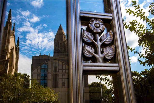 Photo of an ornate metal window grate with a flower design. Marsh Chapel and CAS are reflected in the window. The sky is bright blue and has a few clouds; a tree and other greenery is also reflected.