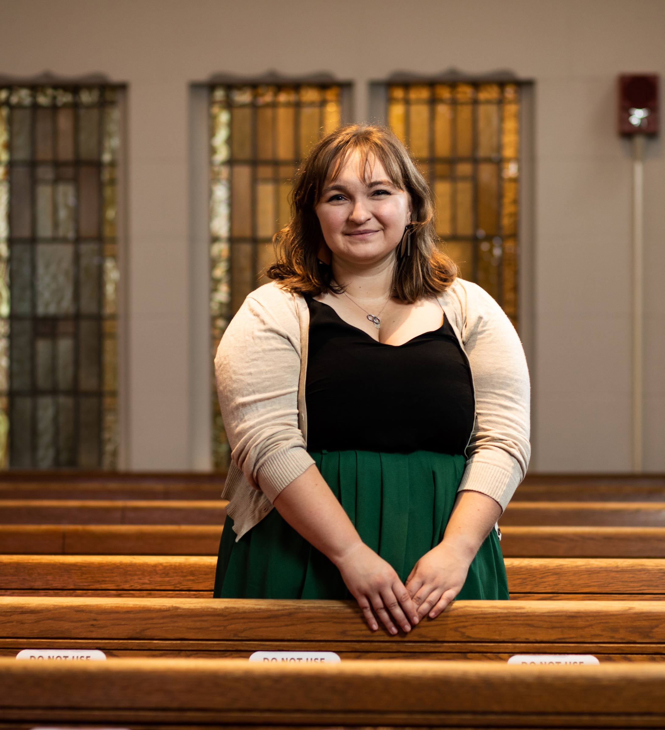 Photo of Jordan Neubauer (LAW’21) in a black and green dress and tan sweater standing between the pews of Marsh Chapel with her hands resting on the pew in front of her. She has dirty blonde shoulder-length hair and smiles.