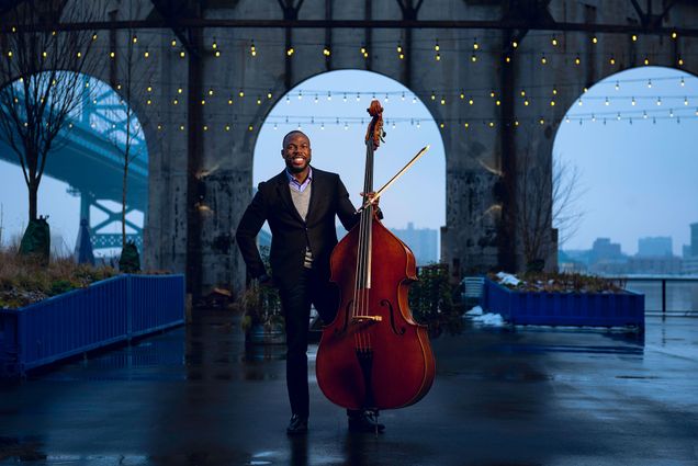Photo of Bassist Joseph Conyers (BUTI’98) in a suit holding his upright bass. He stands in airy, dim room with an arched window in the background, and lights hanging.