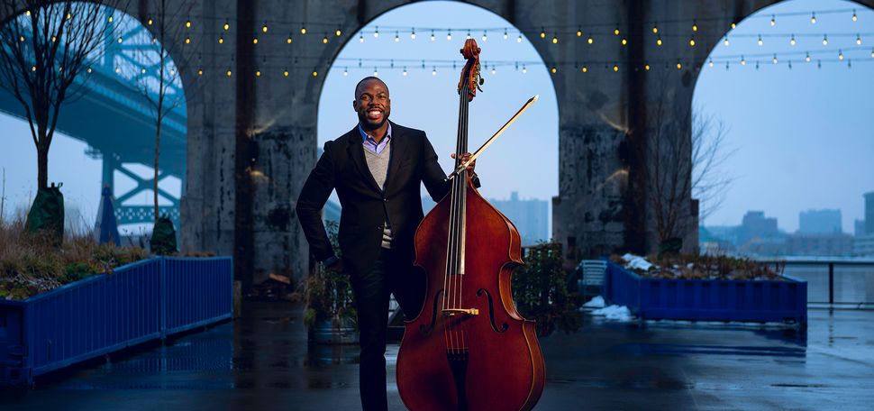 Photo of Bassist Joseph Conyers (BUTI’98) in a suit holding his upright bass. He stands in airy, dim room with an arched window in the background, and lights hanging.