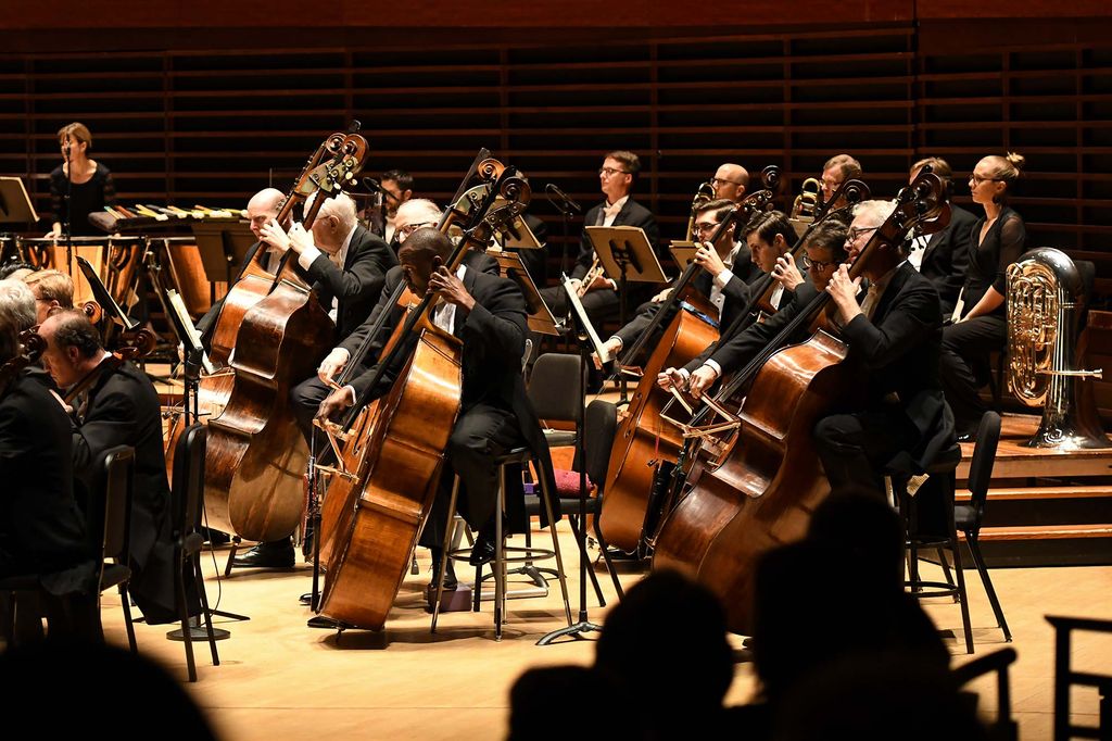 Photo, taken from the right of the stage, as Conyers (center) plays the principal bass in the Philadelphia Orchestra. The bassists sit on black chairs and look forward towards the conductor.