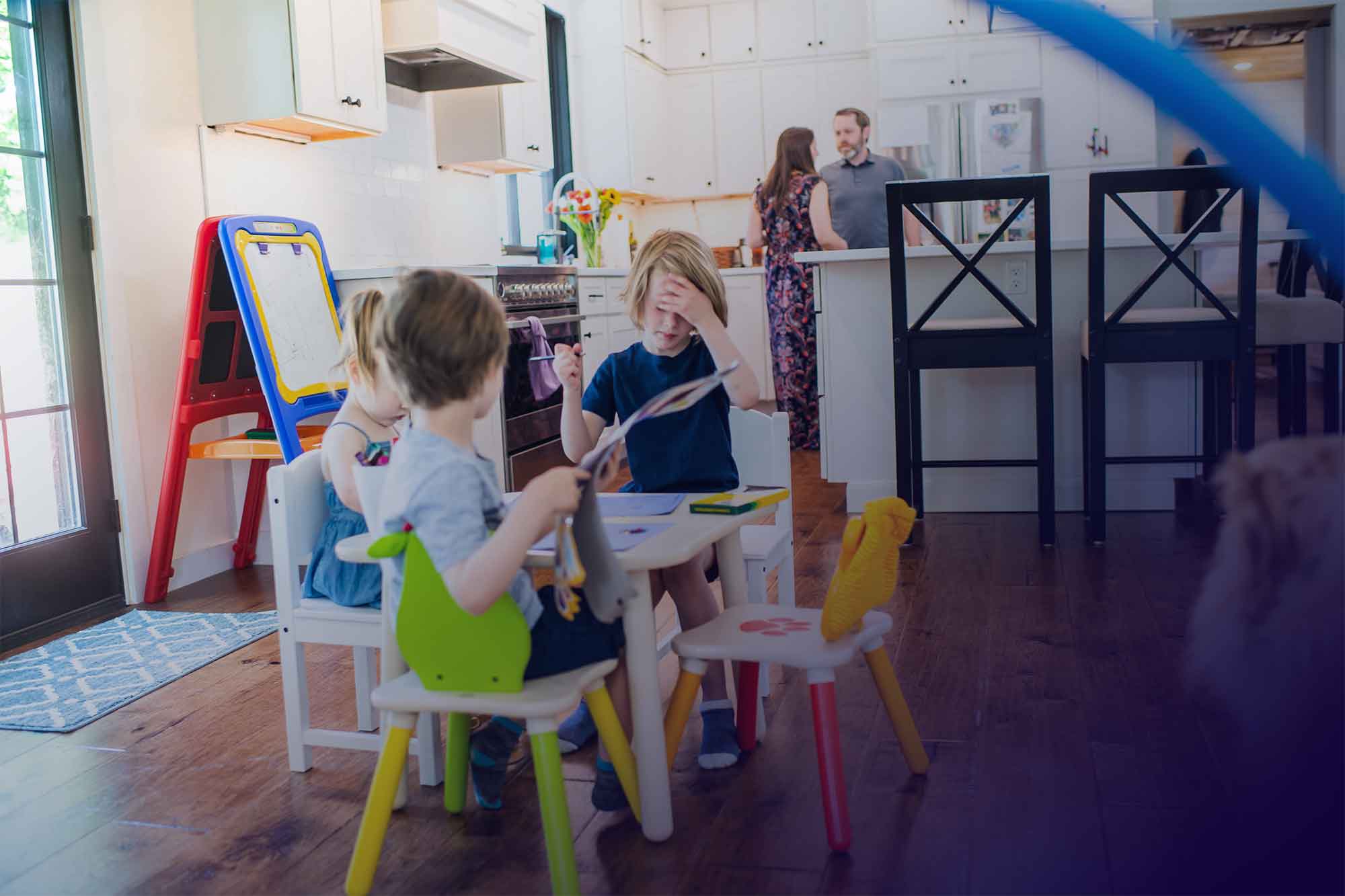 3 children playing or drawing at a kids table while parents talk behind them in the kitchen.